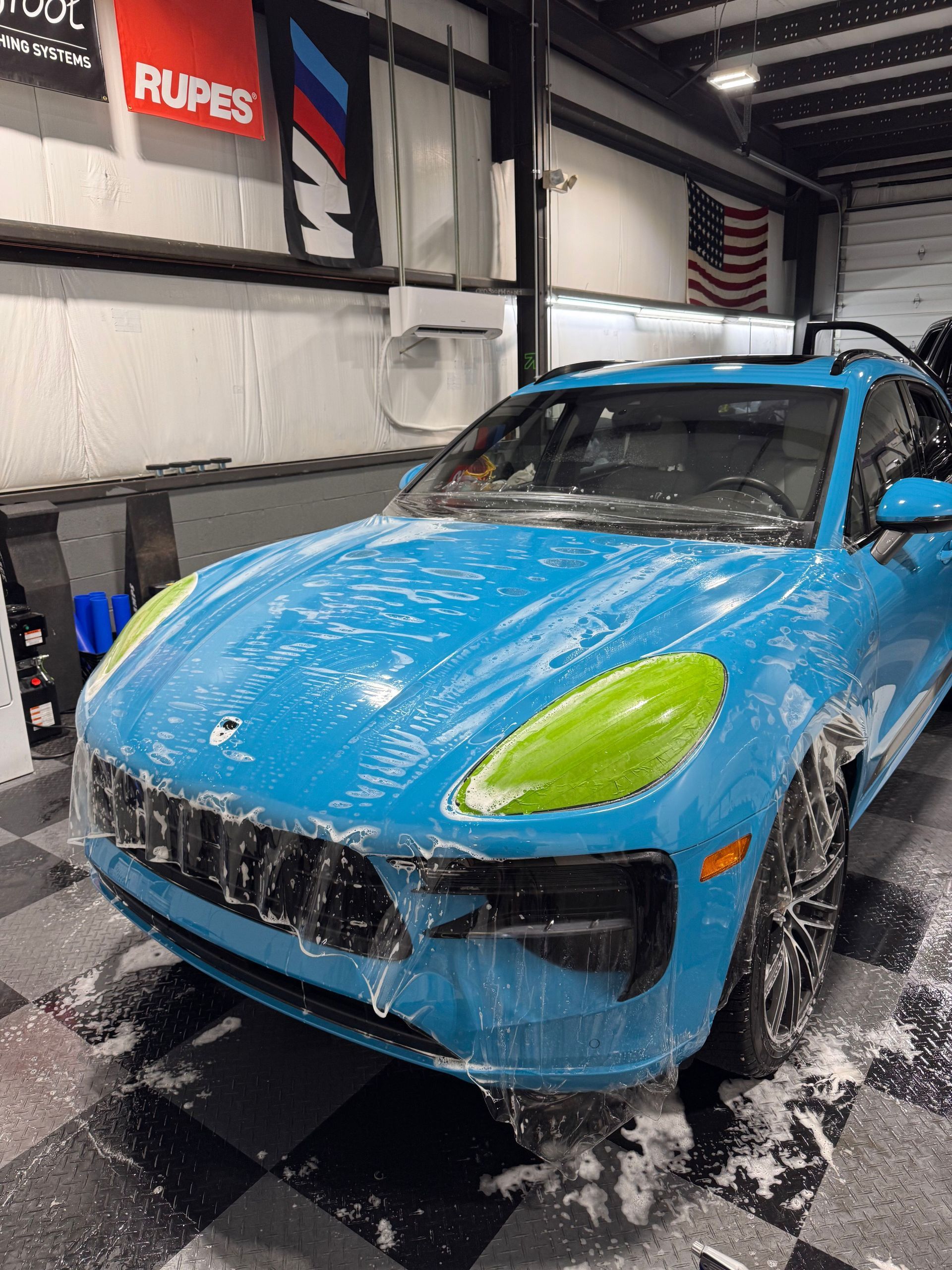 A bright blue Porsche covered in soap suds, parked inside an auto detailing garage with checkered flooring.