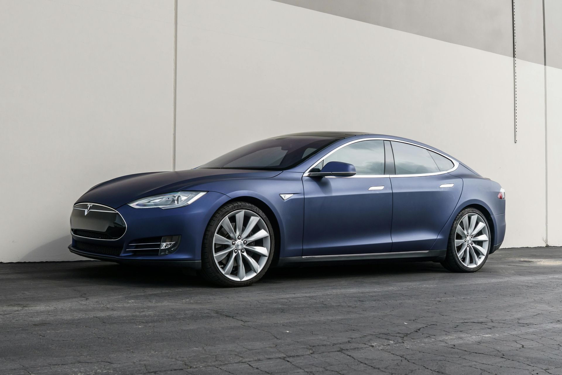 A dark blue Tesla Model S parked on a gray surface against a plain, light-colored wall.