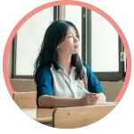 Student sitting at a classroom desk by a window, looking outside with a pen in hand