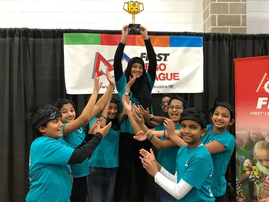 Team in teal shirts lifts a trophy together on a stage with banners in the background.