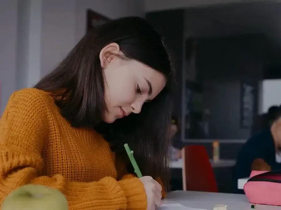 A young woman wearing an orange sweater writing with a green pen