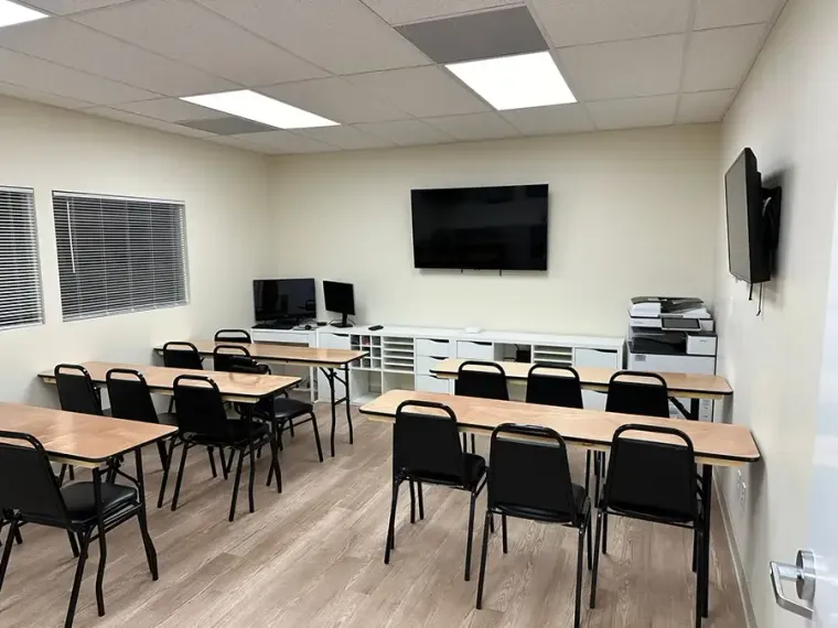 Empty classroom with desks, black chairs, wall-mounted TV, and computer monitors near the front.