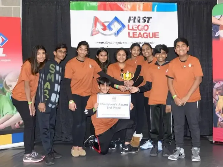 Group of people in orange shirts posing with a trophy at a First Lego League event stage