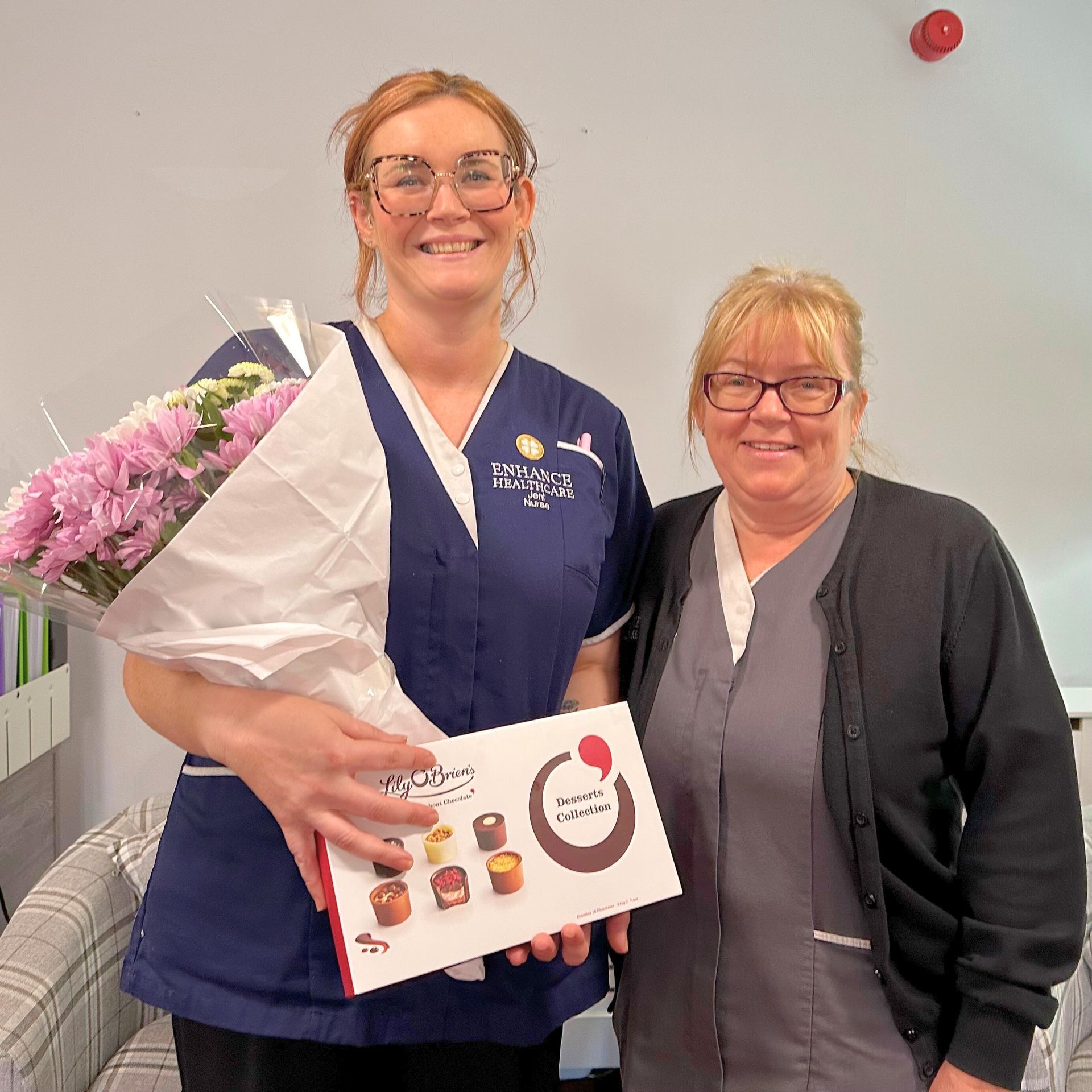 Two women in scrubs pose smiling, one holding flowers and chocolates.