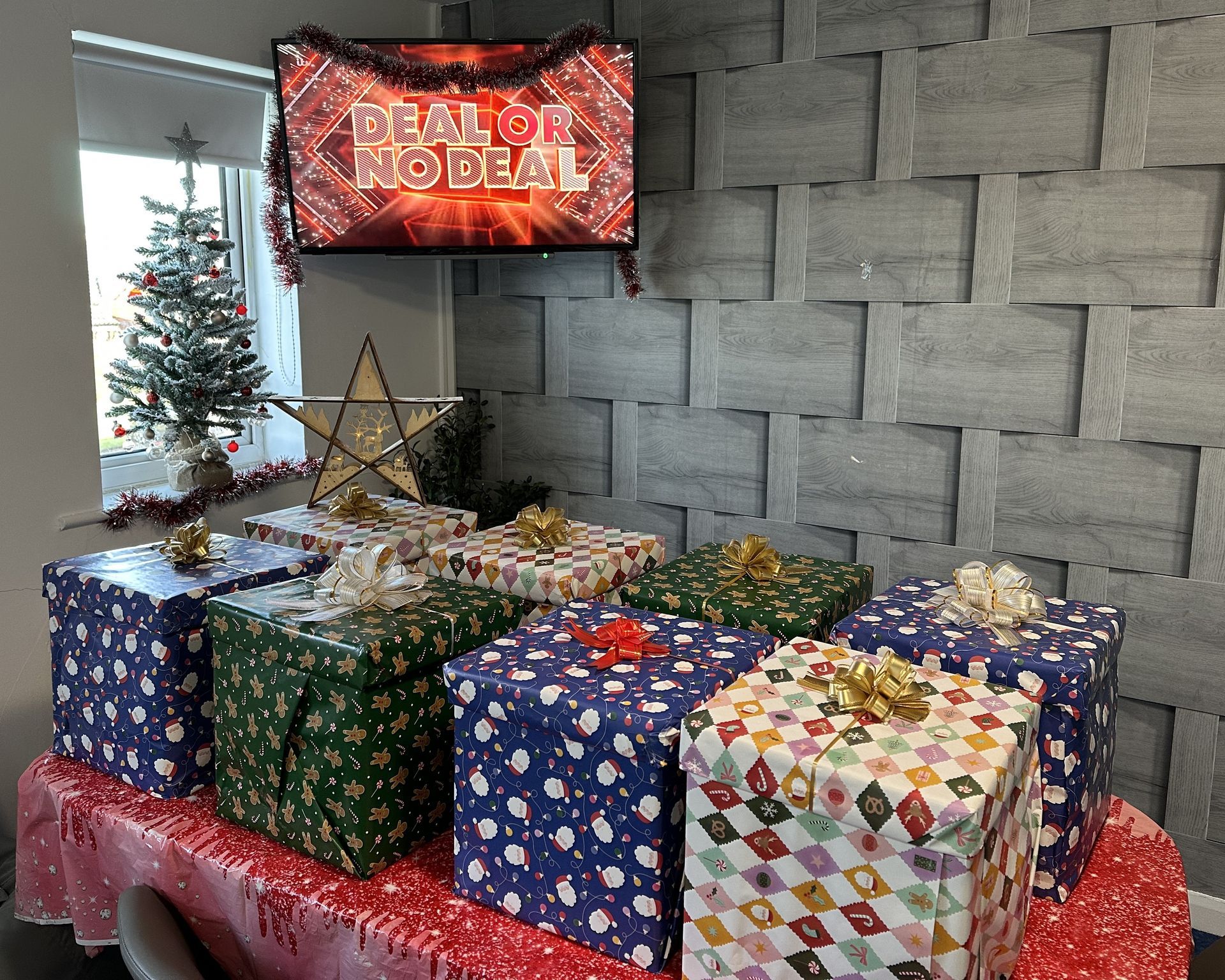 Presents wrapped in festive paper on a table with a holiday sign above. Christmas tree and star in the background.