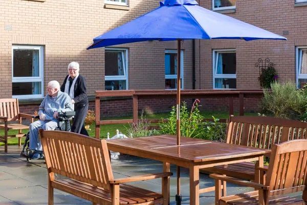 An older person in a wheelchair and another person outdoors, under a blue umbrella near a building.
