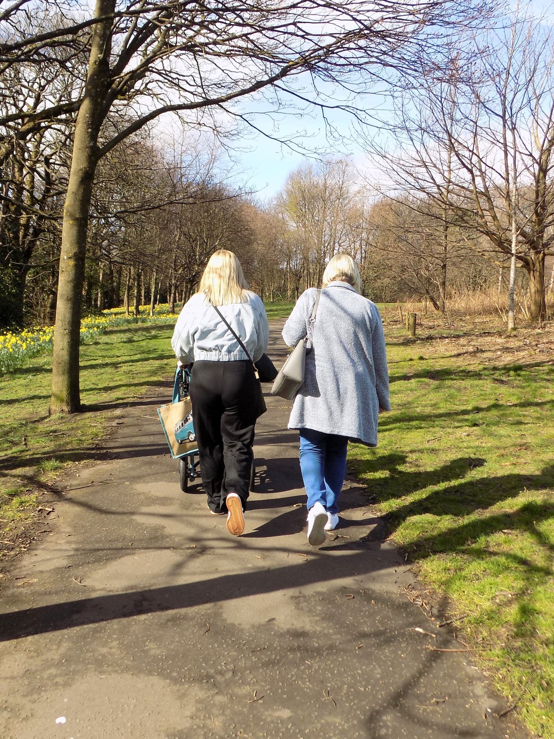 Two women are walking down a path in a park pushing a wheelchair.