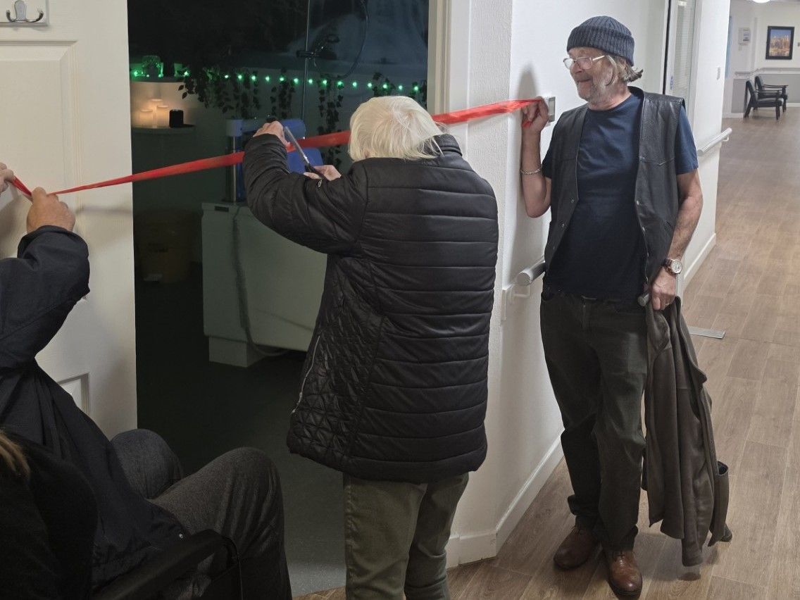 Three people cutting a red ribbon in a doorway. One person sits, another cuts the ribbon, and one smiles.