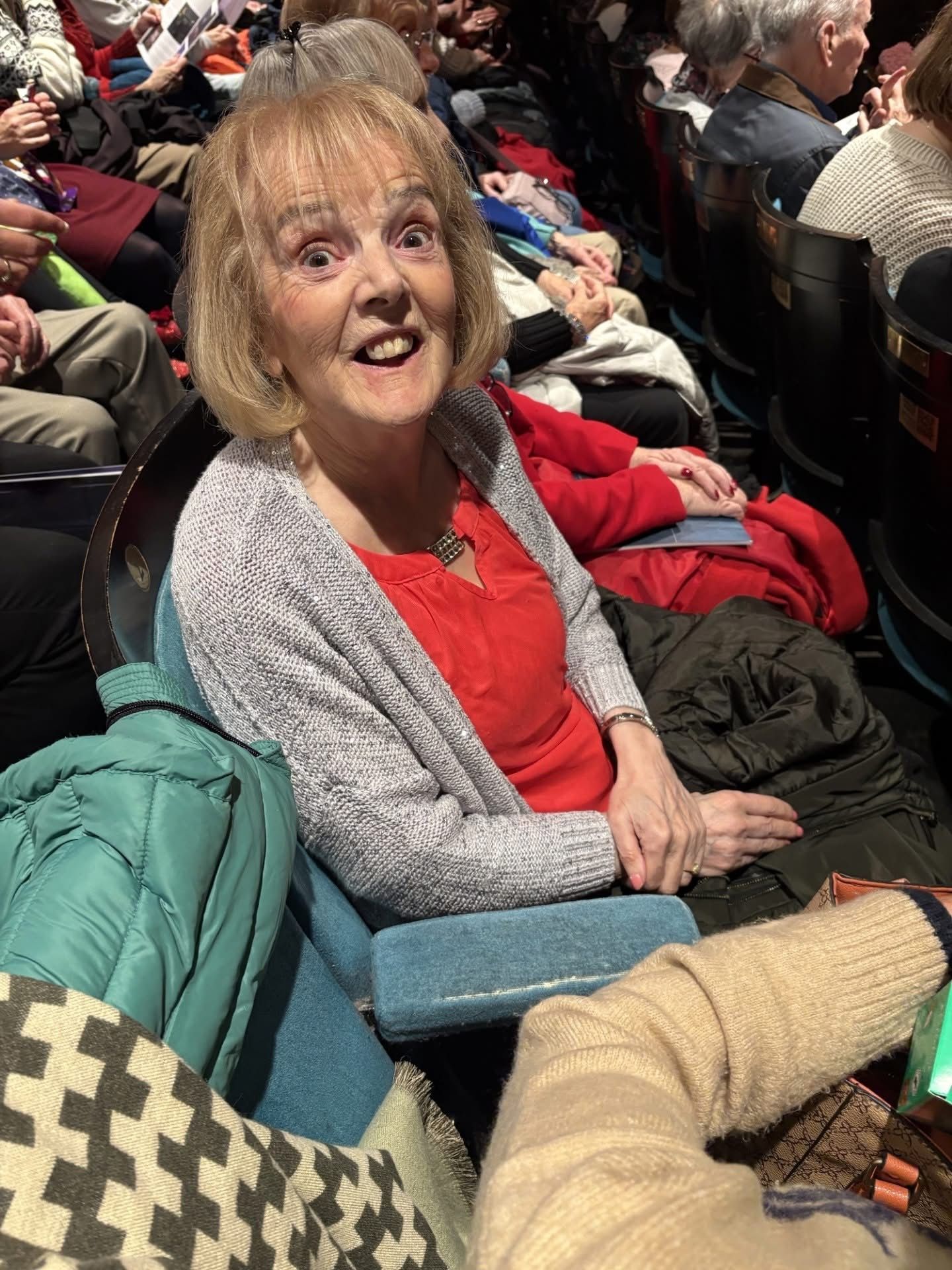 Woman smiling in theater seat, wearing red top and gray sweater. Surrounded by other theater-goers.