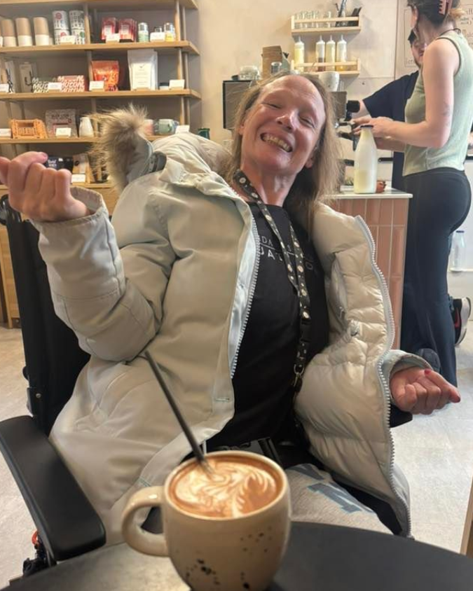 Person in wheelchair smiles, extends arms, holds coffee with latte art. Inside a cafe.