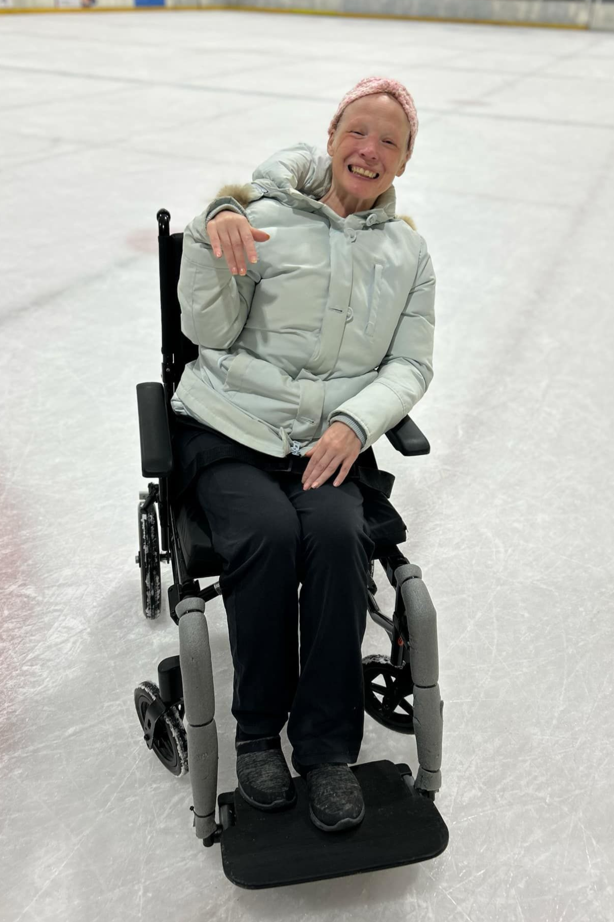 Woman in wheelchair smiling on an ice rink, wearing a light-colored jacket and a knit cap.