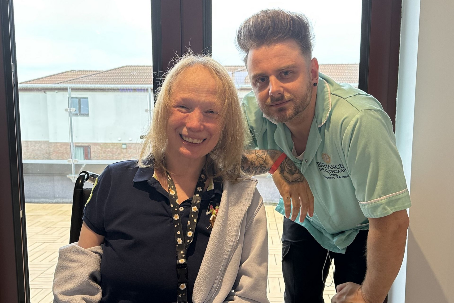 Woman in wheelchair smiles with man, both in front of a window.