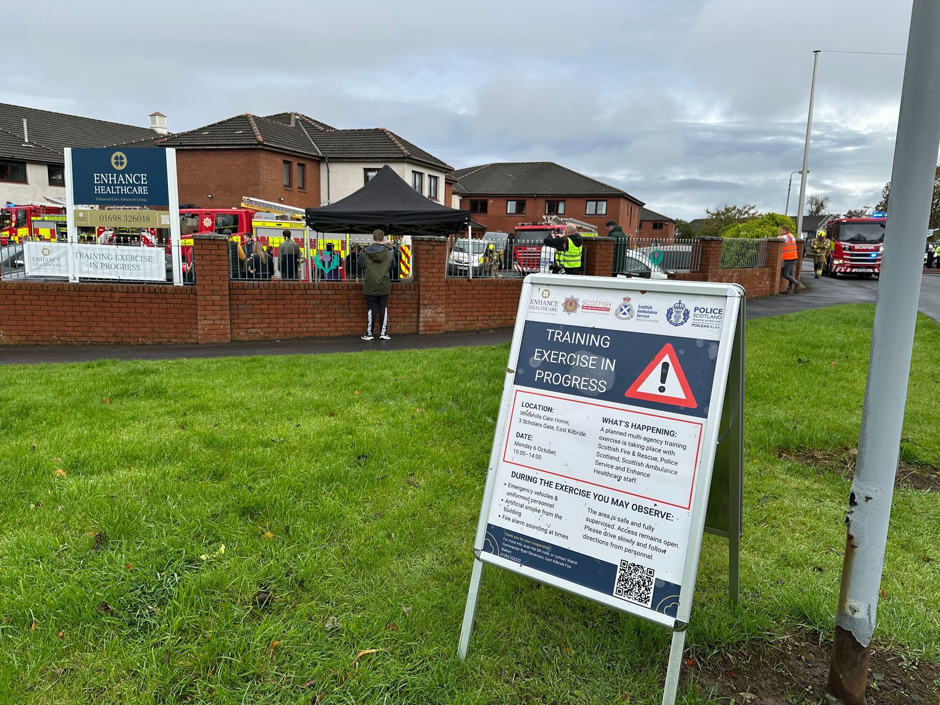Sign in front of building with fire engines. Warning sign. People gathering, overcast.