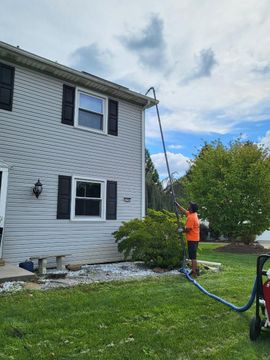 Worker in orange shirt cleaning white house siding with a pressure washer in a yard.