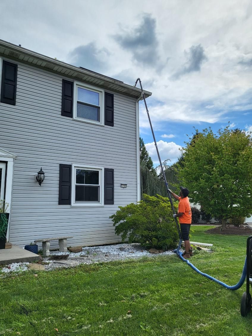 Worker in orange shirt pressure-washing the side of a white house with black shutters.