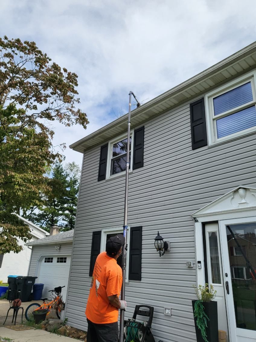 Person in orange shirt standing beside a gray two-story house with a ladder leaning against it.