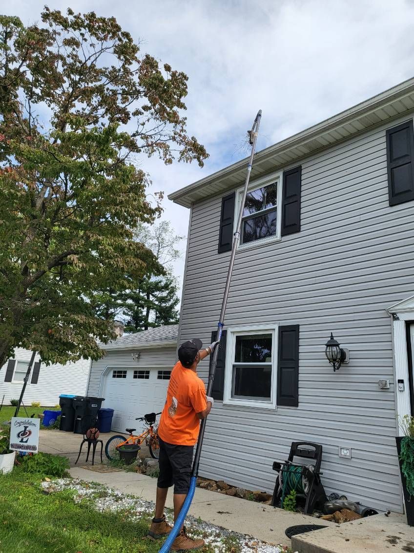 Person in orange shirt painting the exterior of a two-story white house with a roller on an extension pole.