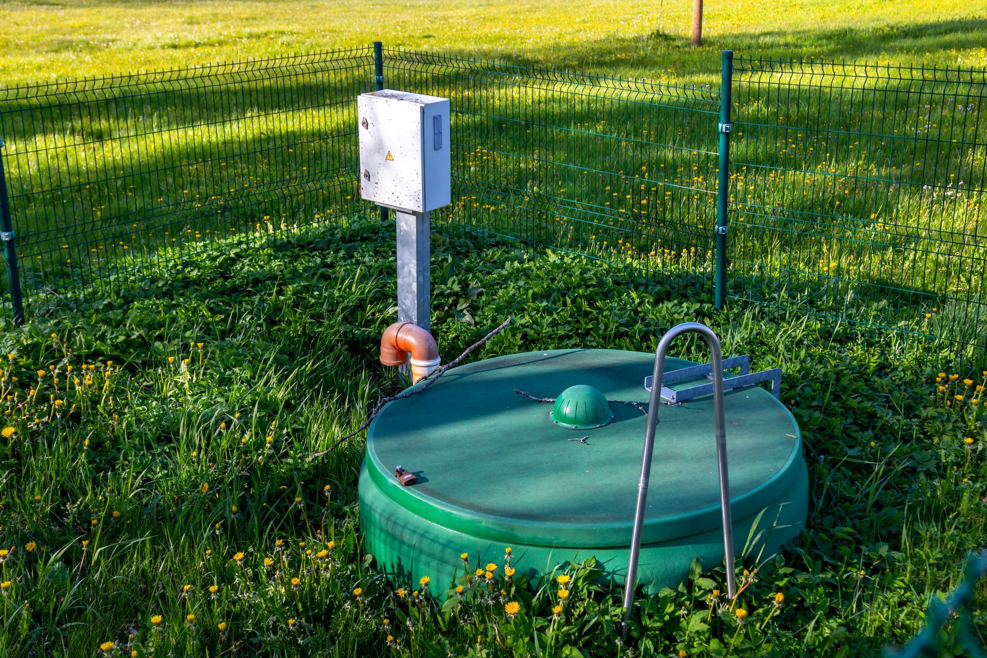 A green, circular utility cover sits in a grassy field next to a white control box on a post.