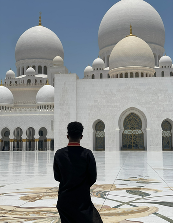 Person in black stands in a white mosque courtyard with large domes and arched windows.