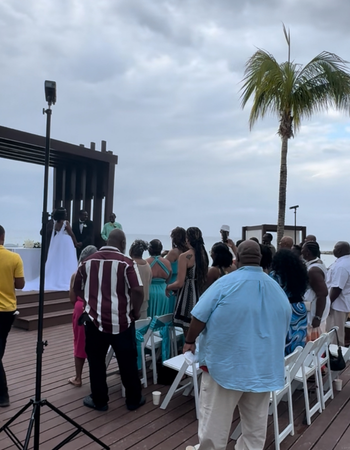 People seated on a wooden deck facing a stage outdoors under a cloudy sky, with a palm tree nearby