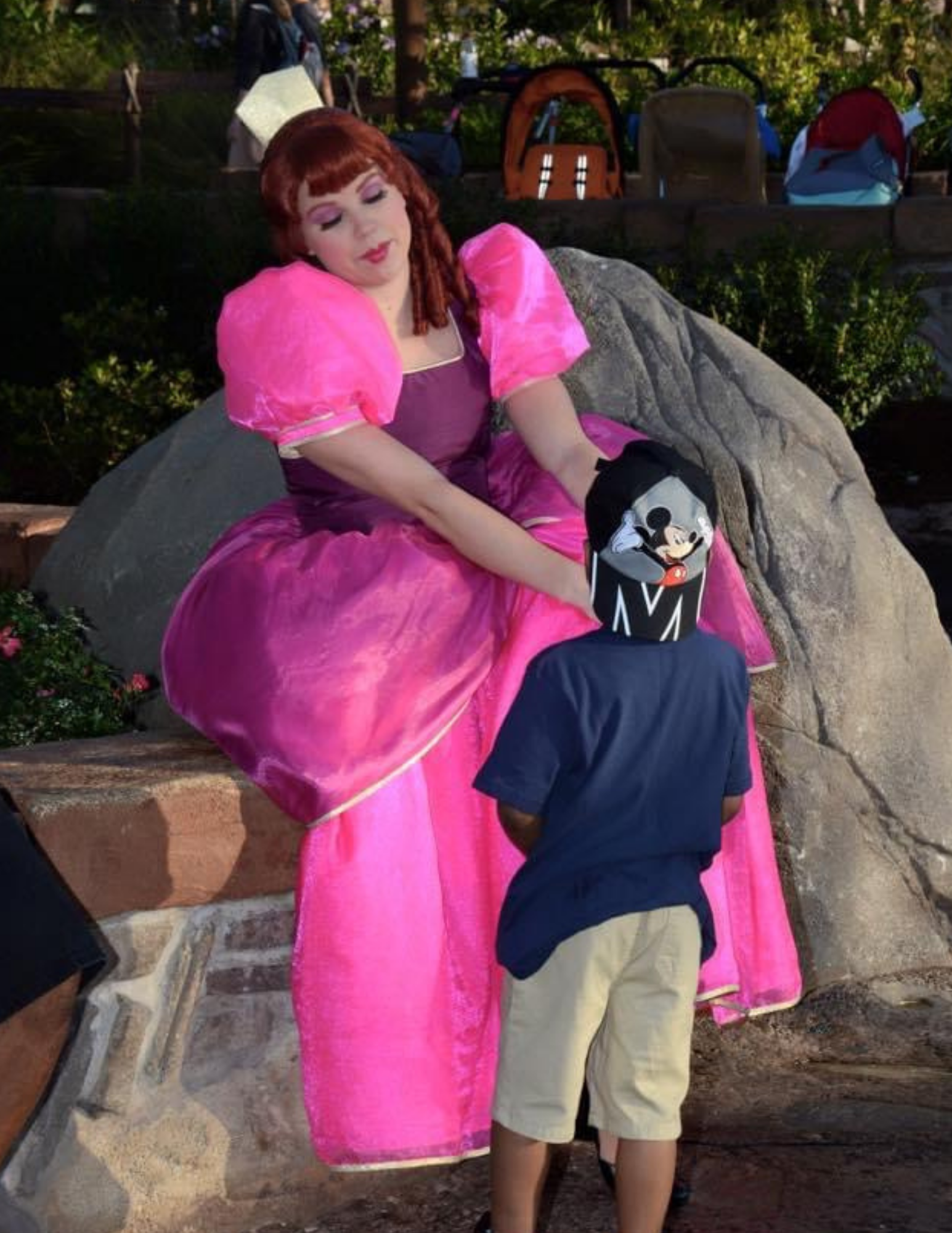 Woman in bright pink dress kneels by a child in a toy hat beside a large rock outdoors