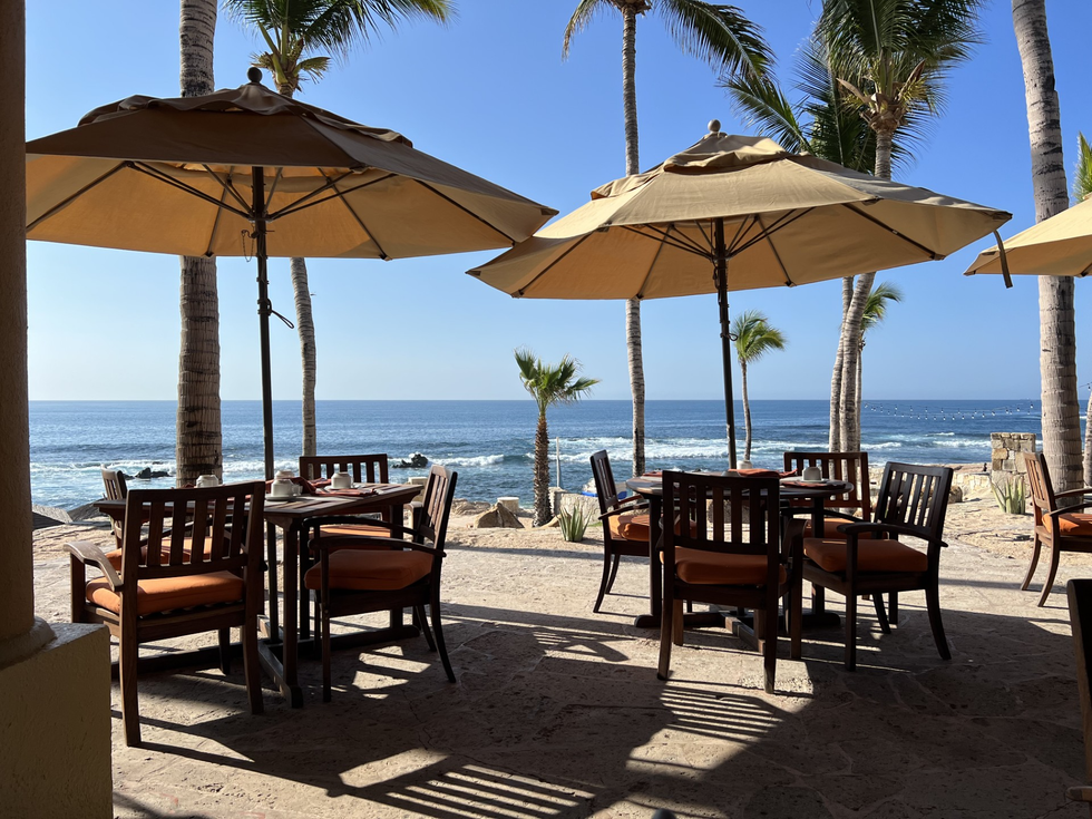 Beachfront patio with wooden tables and tan umbrellas, facing the ocean and palm trees under a clear blue sky