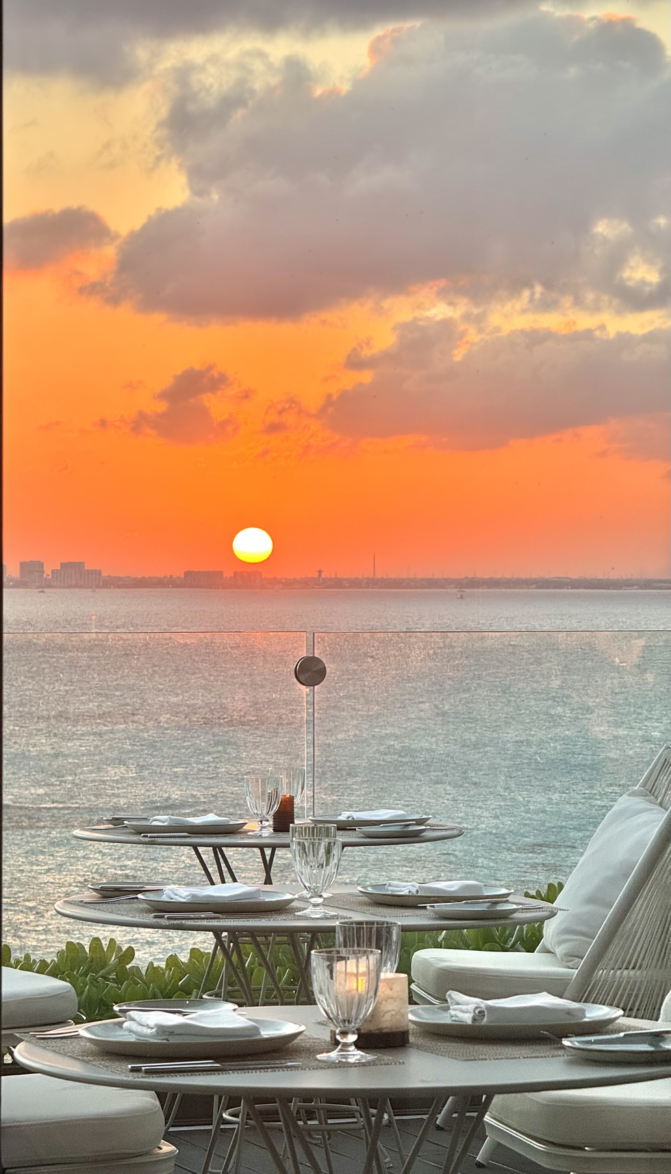 Sunset over calm ocean seen from a seaside dining table with candles and white chairs