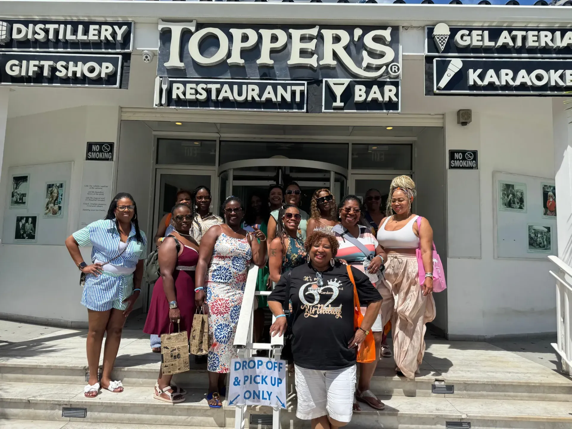 Group posing outside Toppers restaurant and bar, with signs for gelateria, distillery, gift shop, and karaoke.