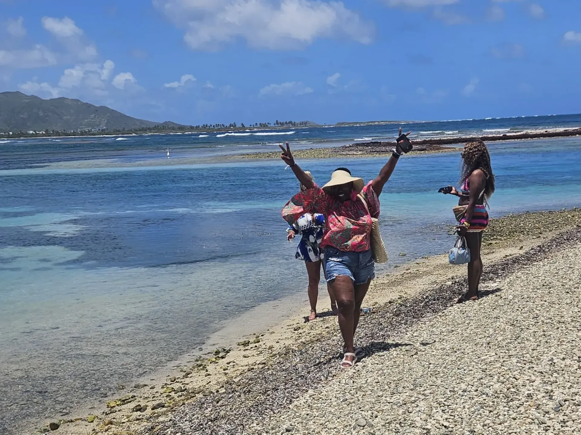 Three people in life jackets stand on a pebbly beach by clear blue water, one with arms raised.