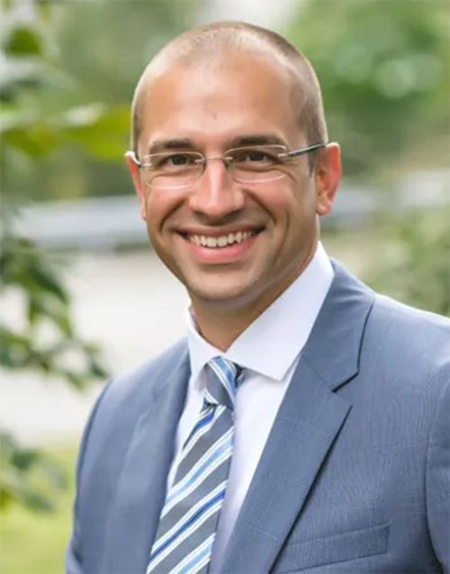 Smiling man in a blue suit and striped tie outdoors with a blurred green background