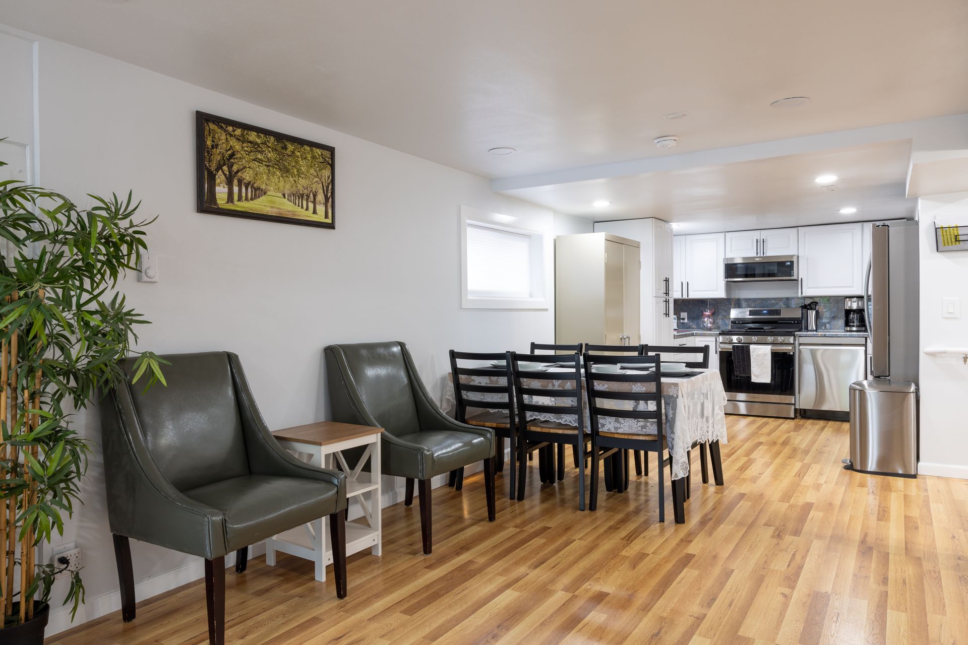 Bright living space with dining table, chairs, and kitchen in the background. Wood floors and neutral walls.