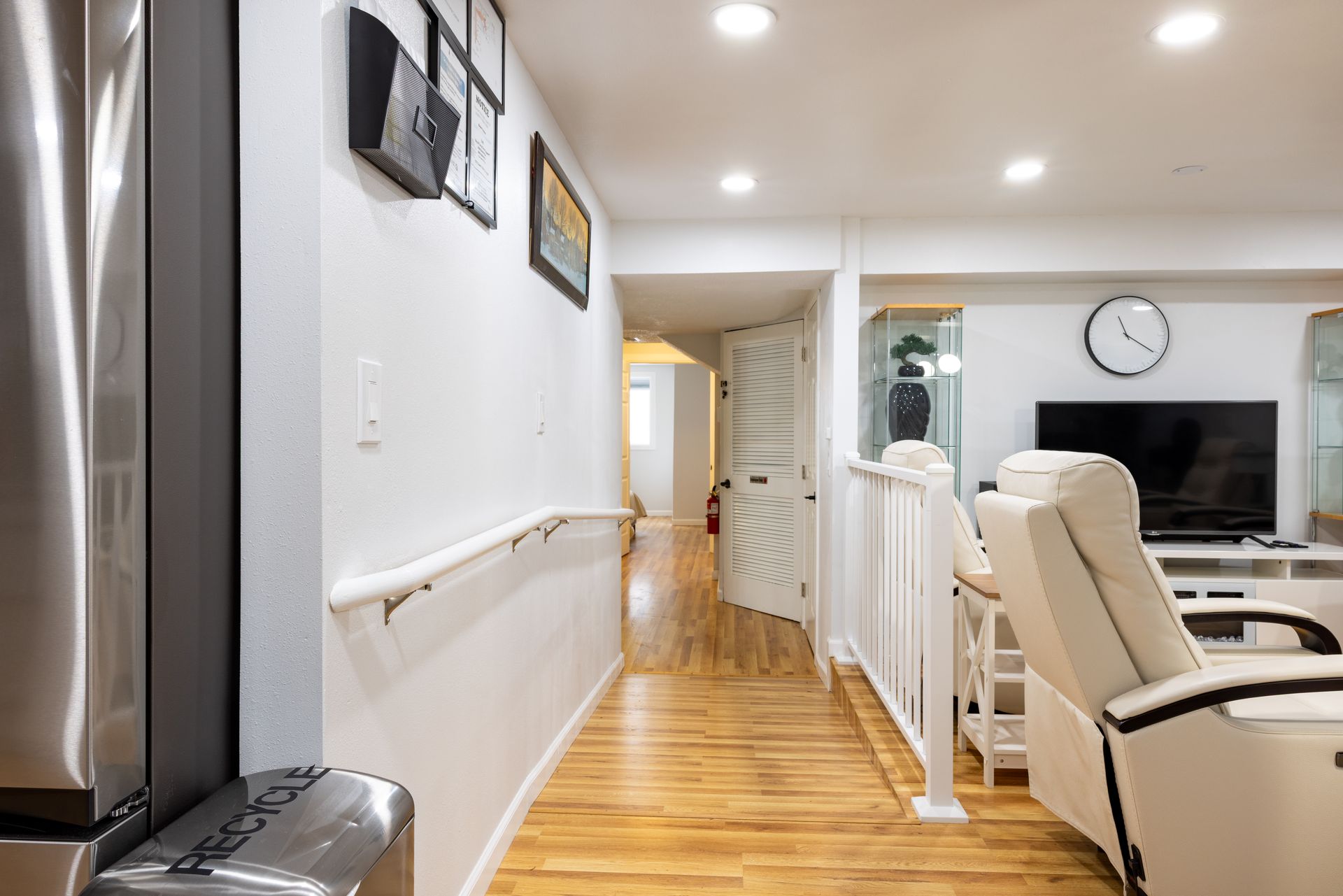 Interior hallway with hardwood floors, handrail, and white walls leading to a door; recliner in the foreground.