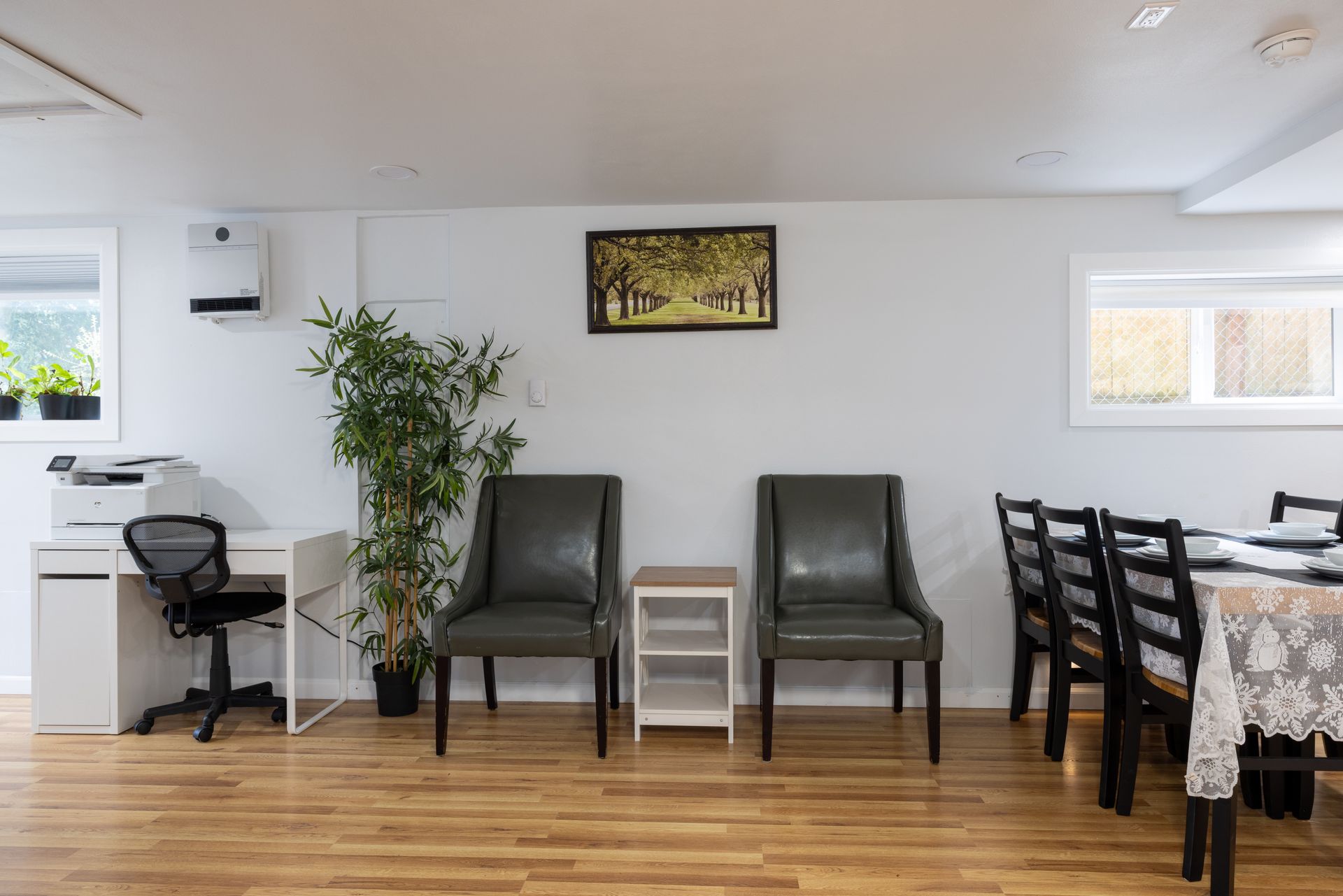 Room with a desk, chairs, and table. Features include a plant and artwork against a white wall and a wooden floor.