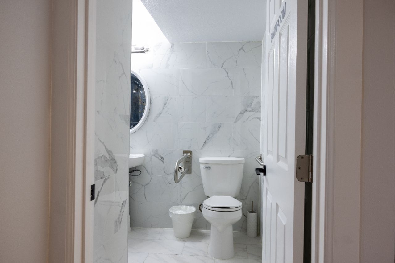 Small, white tiled bathroom with a toilet, sink, and mirror, viewed from the doorway.