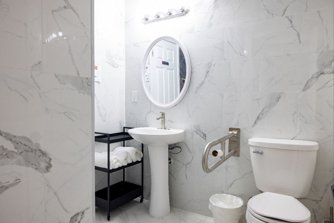 White-tiled bathroom with pedestal sink, toilet, grab bar, and round mirror. A small black shelf holds towels.