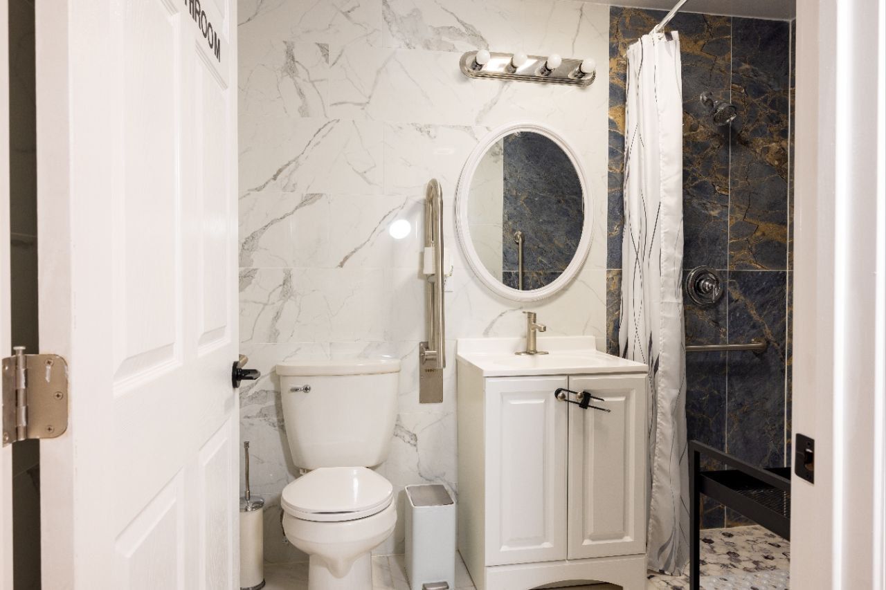 Bathroom with marble-patterned walls, toilet, vanity, oval mirror, shower, and safety grab bar.