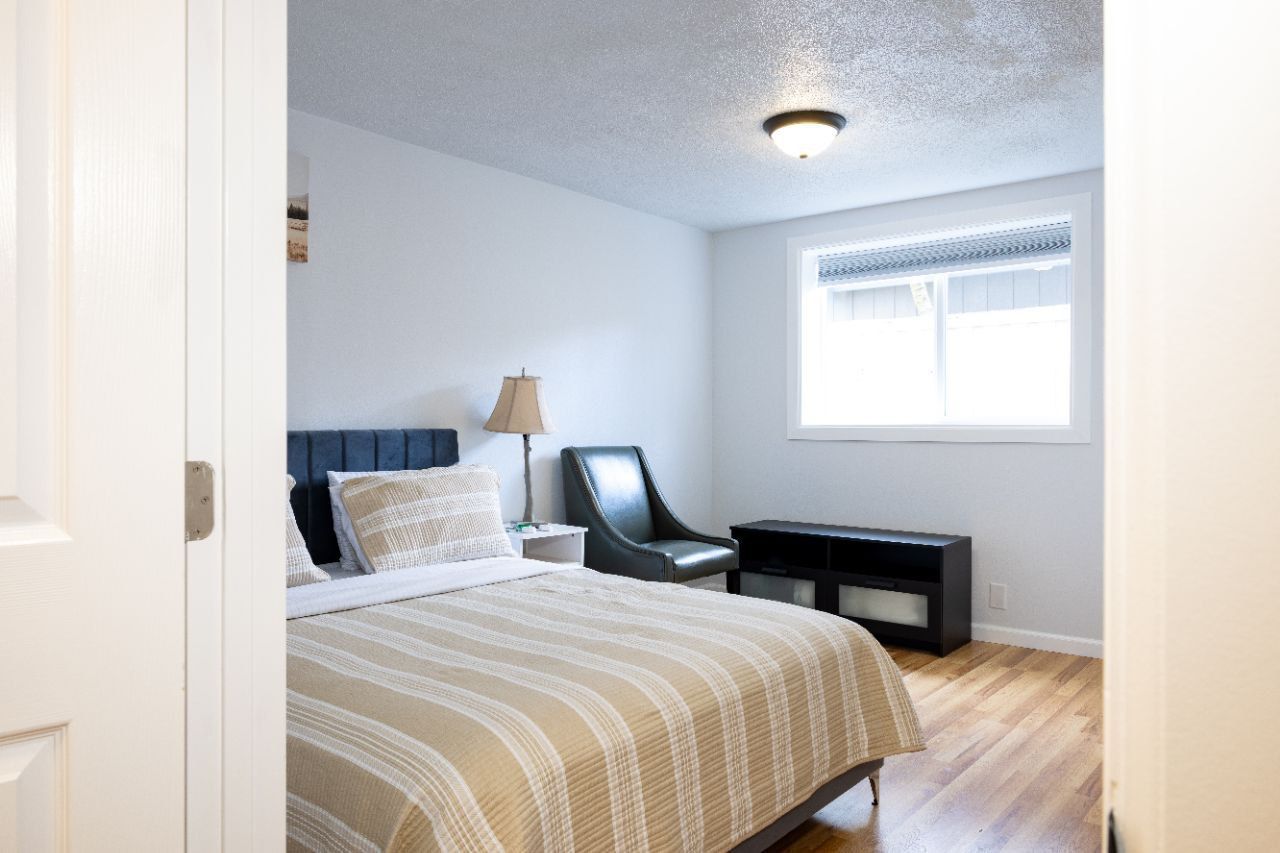 Bedroom interior: bed, window, chair, nightstand. Light wood floors, white walls, and a recessed ceiling light.