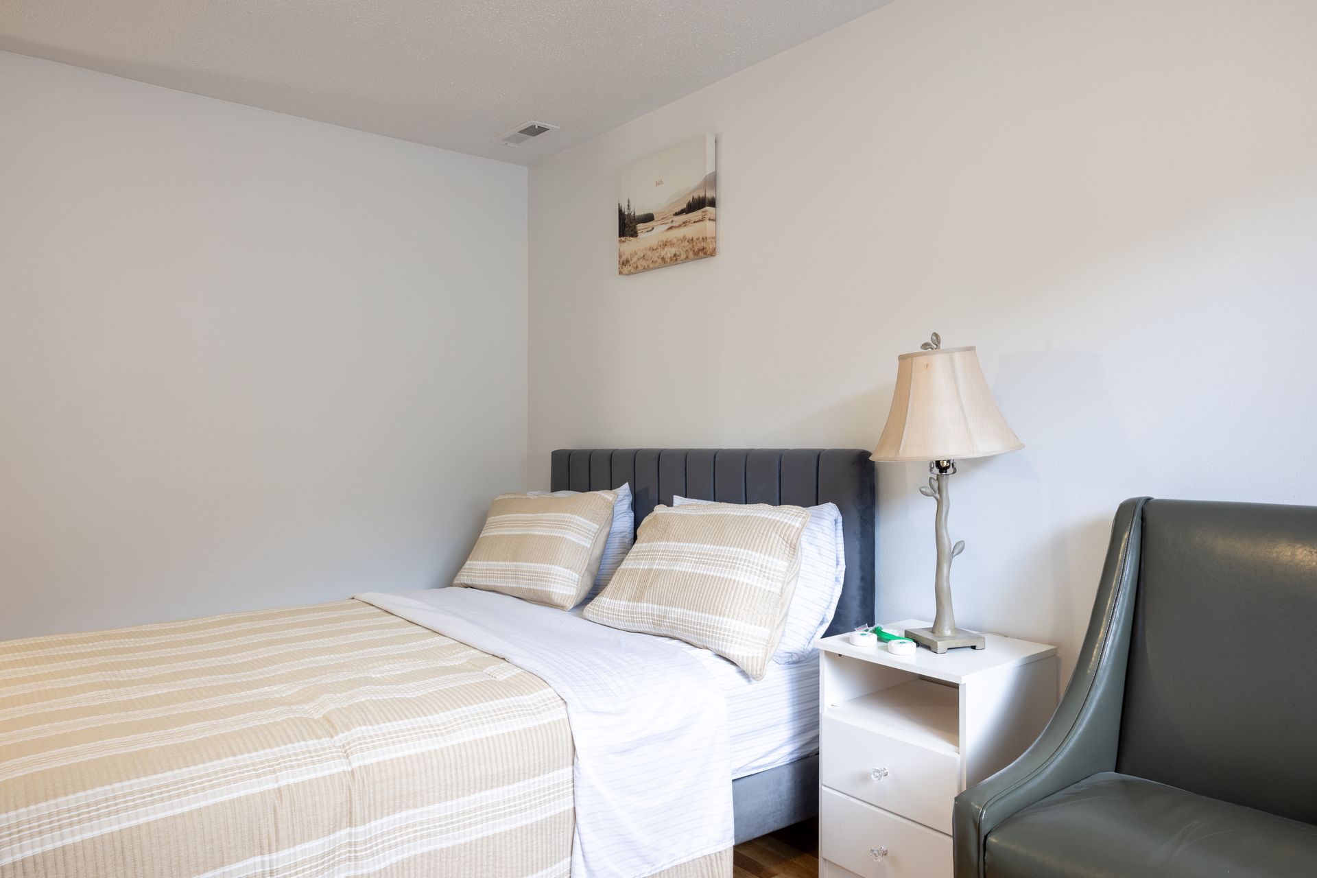 Bedroom with bed, nightstand, lamp, and armchair. Light colored walls.
