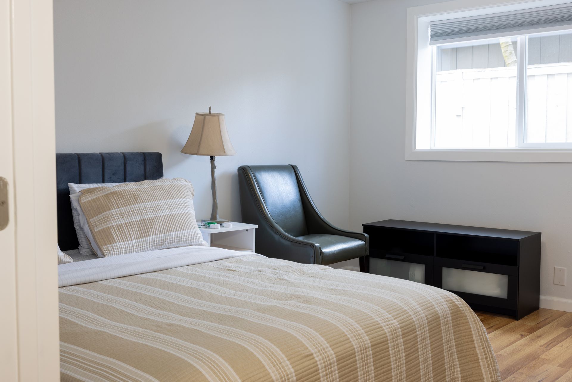 Bedroom with bed, chair, lamp, and window, with light gray walls and wooden floors.