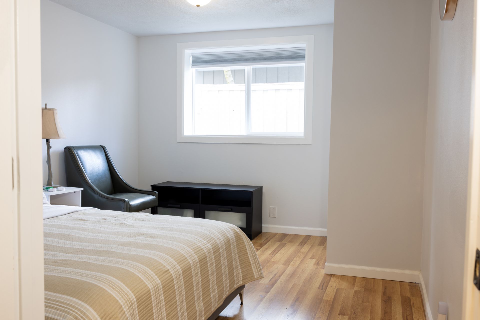 Bedroom with bed, chair, and window. Hardwood floor and neutral-colored walls.