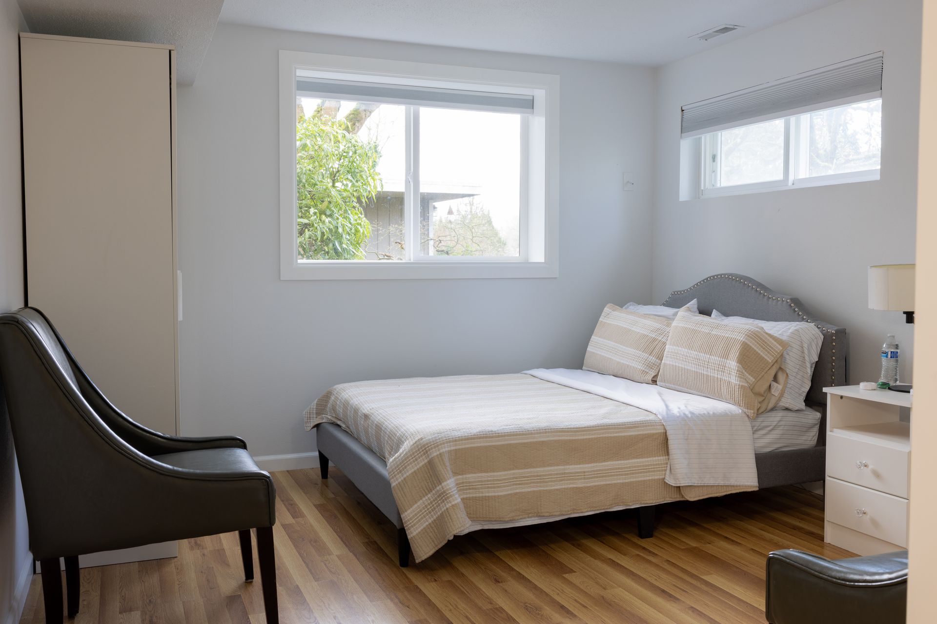 Bedroom with bed, chair, window, closet. Light-colored walls, wooden floor, natural light.