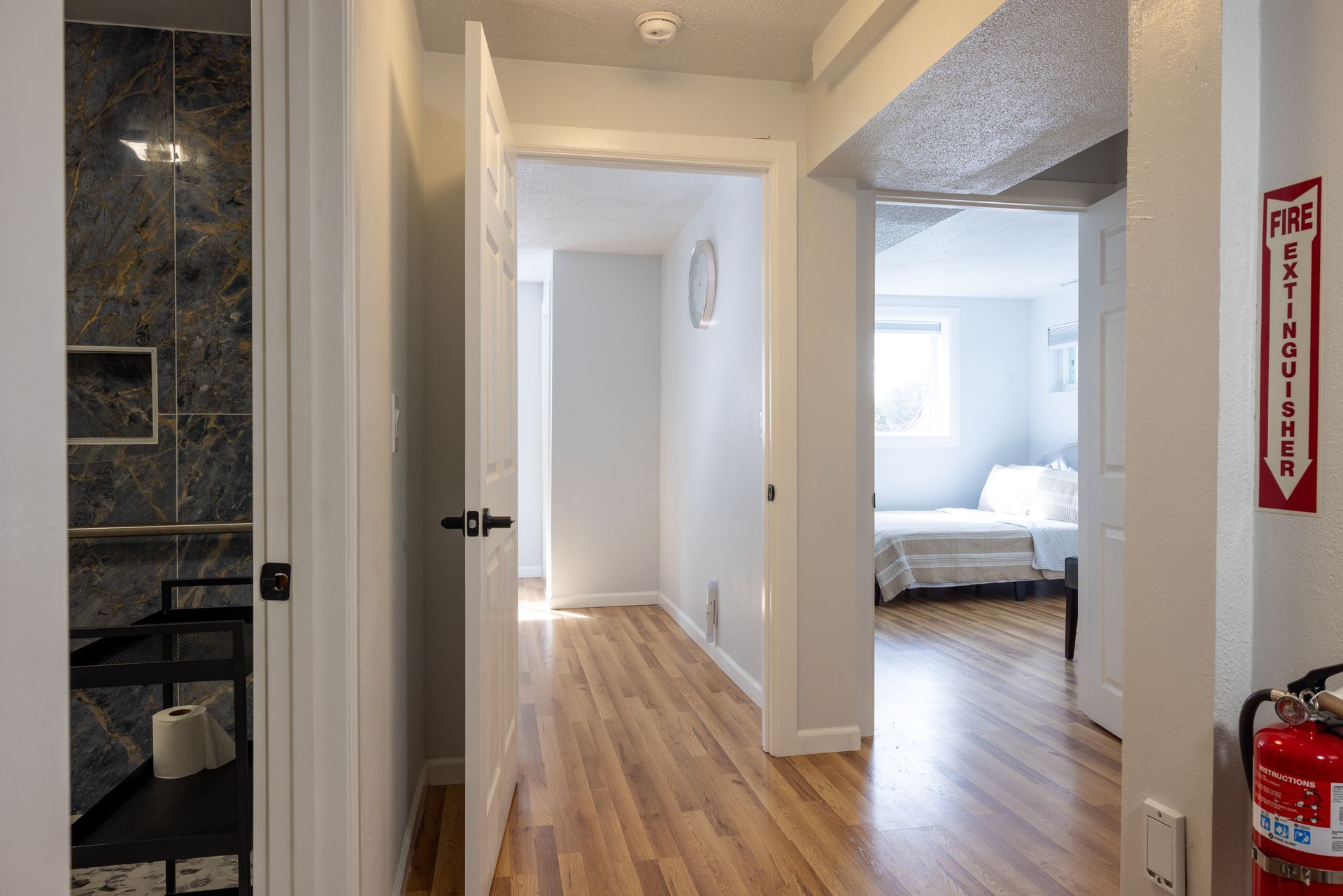 Hallway with hardwood floor, leading to bedrooms. One door open to a bathroom with a tiled shower. Fire extinguisher.