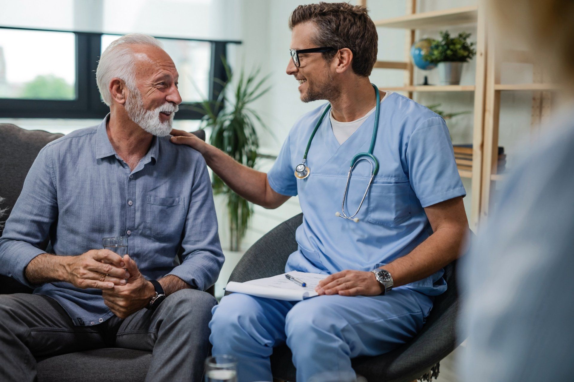 Healthcare provider comforts an older person, indoors. Provider in blue scrubs has arm on patient's shoulder.