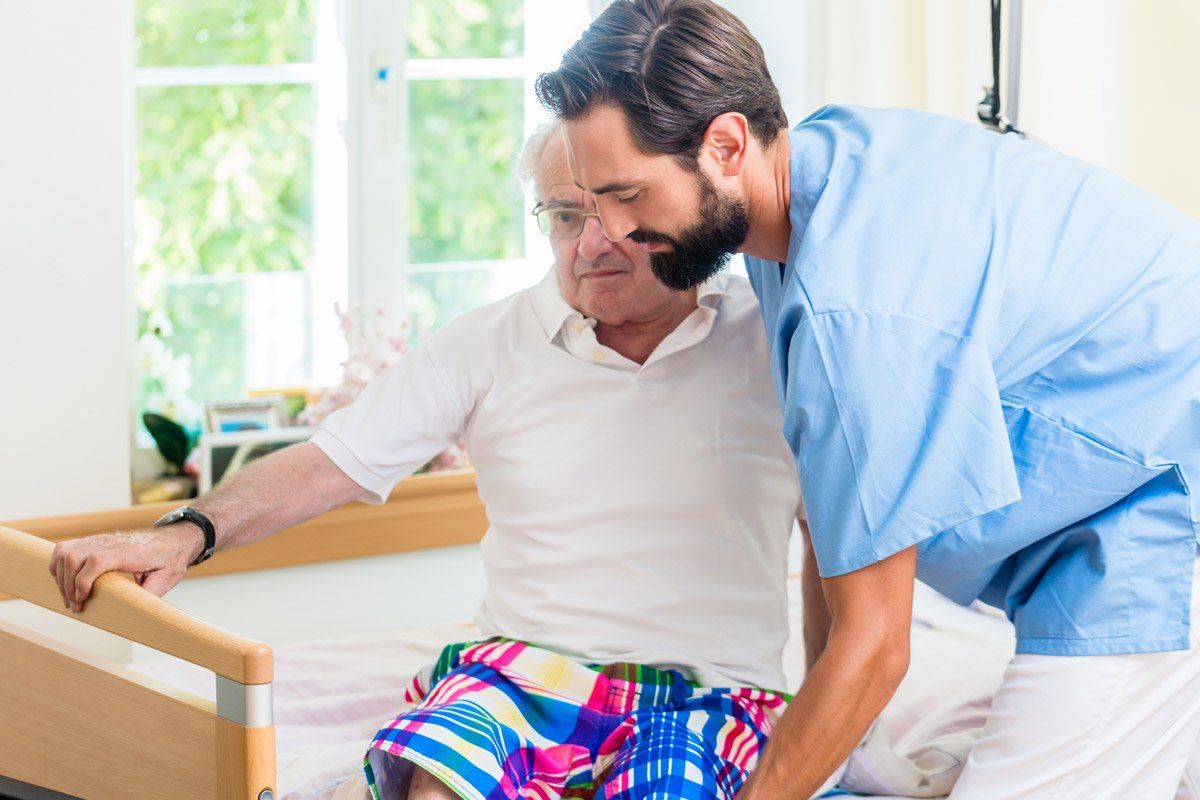 Caregiver assisting a patient out of bed. The patient sits, holding bed rail. The caregiver is supporting. Bright room.