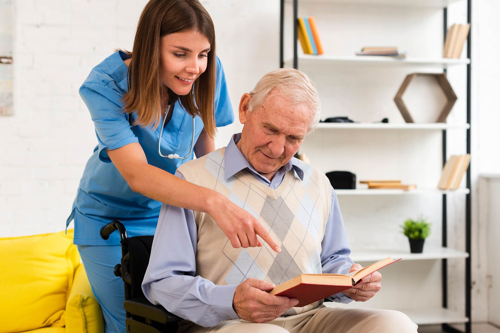 Nurse pointing at book held by older adult in a wheelchair, indoors.