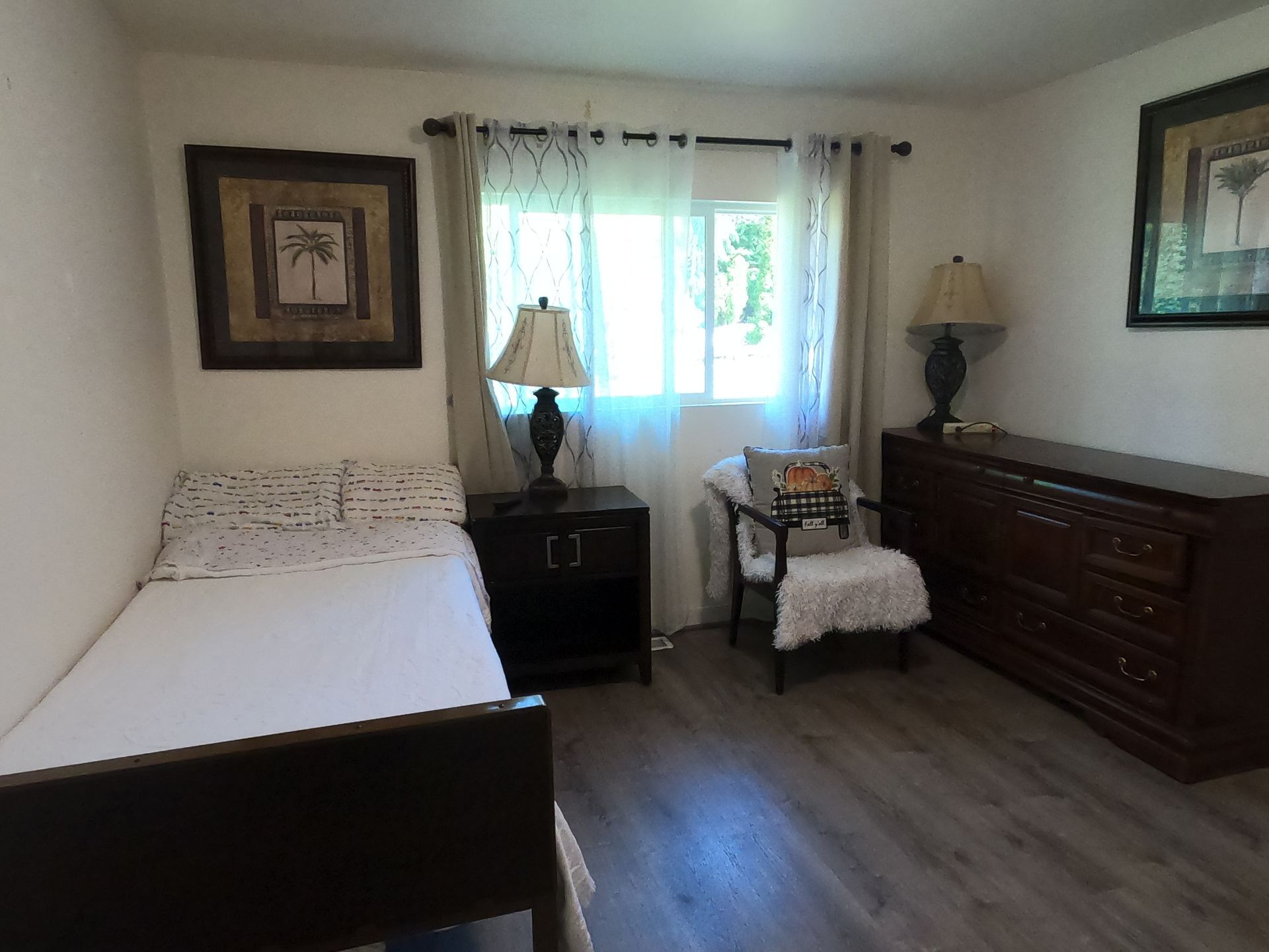 Bedroom with single bed, nightstand, dresser, armchair, window with curtains, artwork, and lamps.