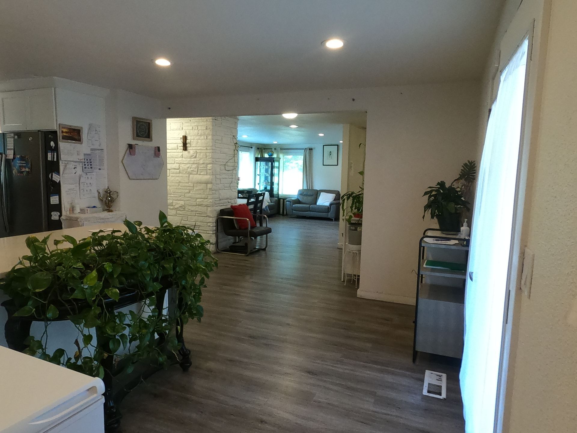Interior view of a home with open doorways. Plants and a refrigerator are visible. Grey flooring and white walls.