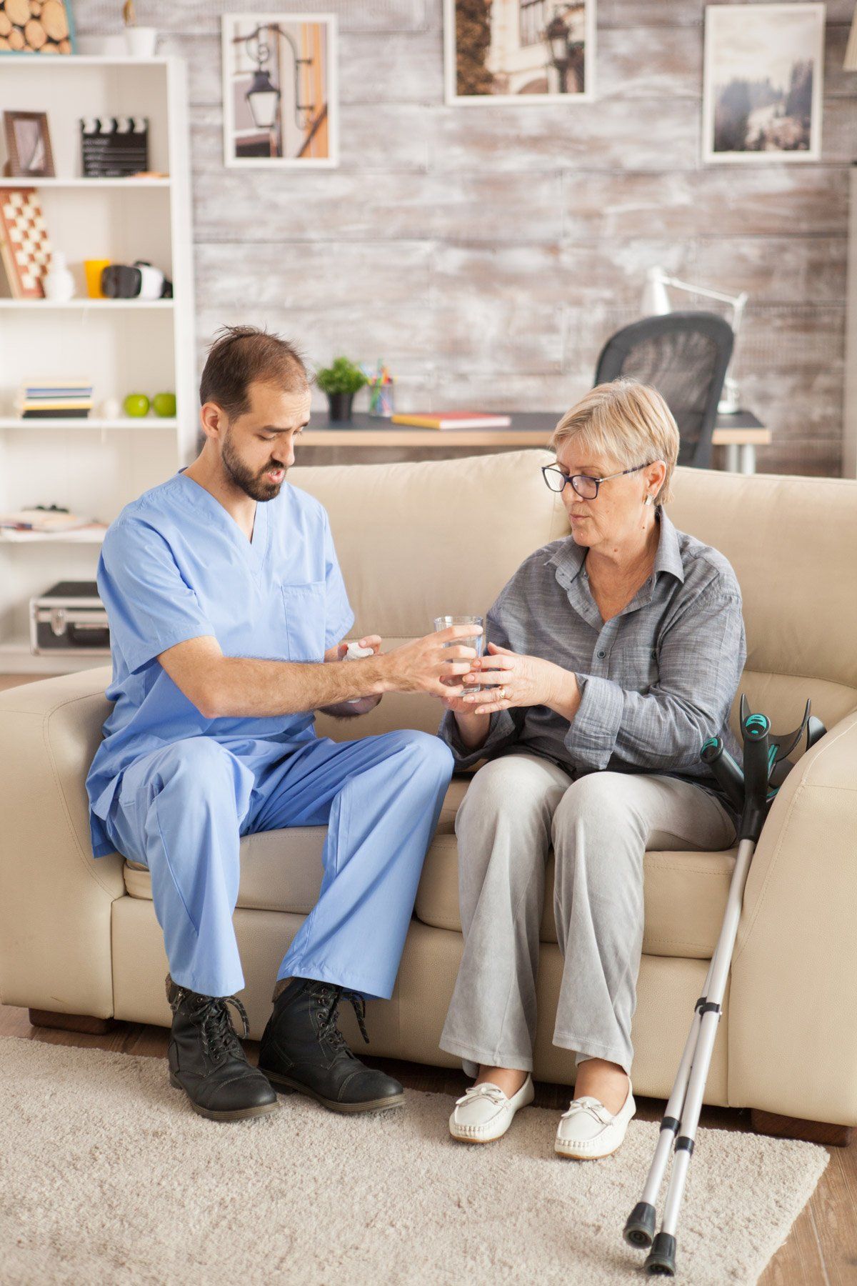 Caregiver offers a glass of water to an older person seated on a sofa indoors. A crutch rests beside them.