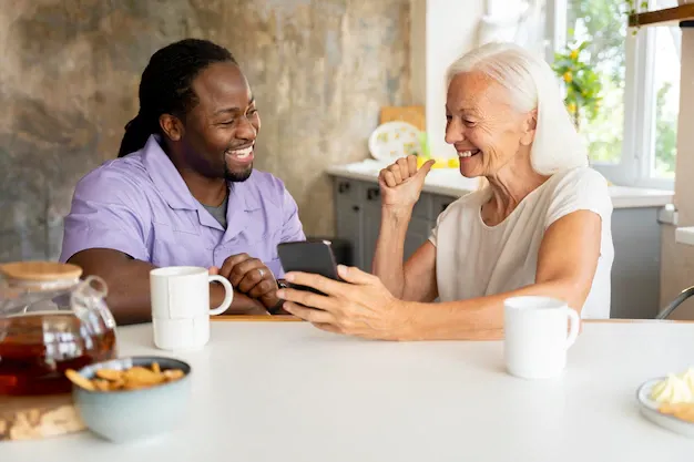 Man and woman smiling, looking at a phone, sitting at a table in a kitchen.
