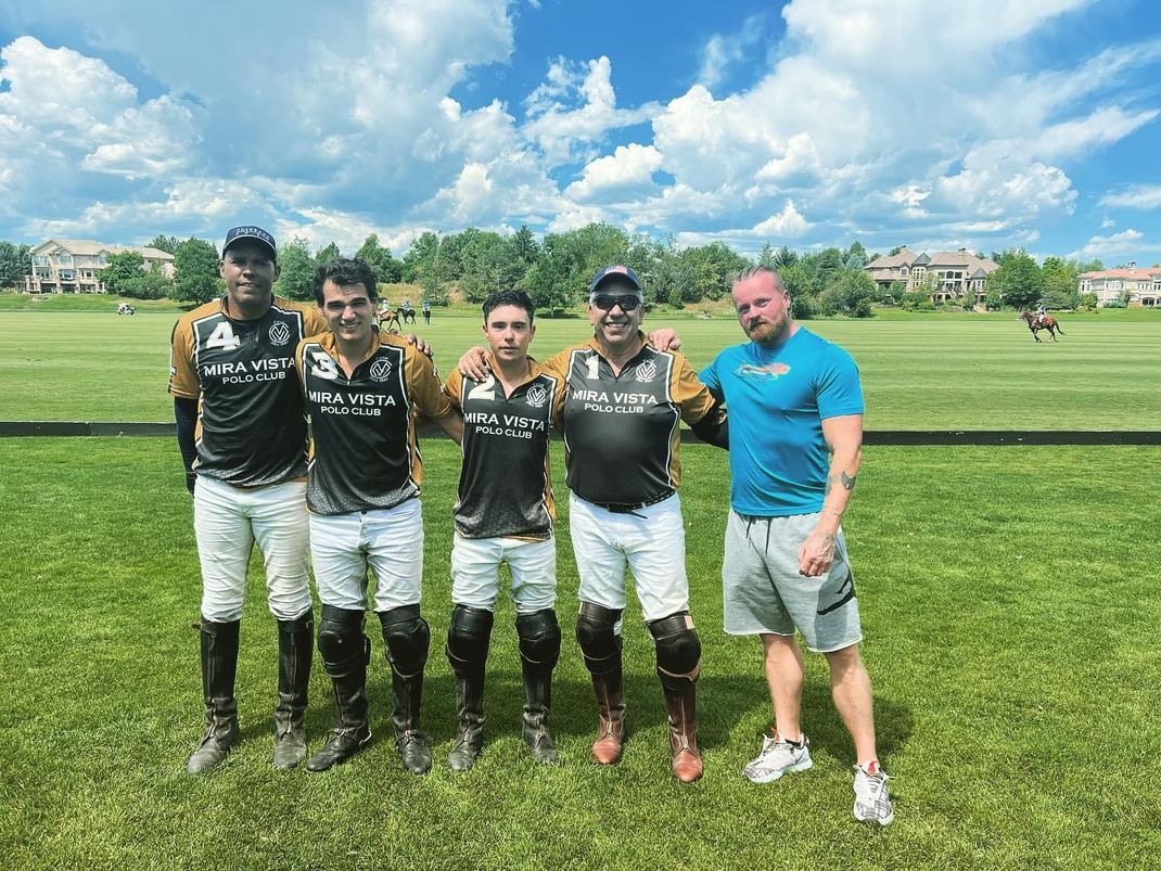 Four polo players on horseback line up on a grassy field under a clear blue sky, holding mallets.